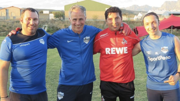 Three coaches of the Young Bafana Soccer Academy and one coach of the 1. FC Cologne standing arm in arm on the pitch of the YB Arena.