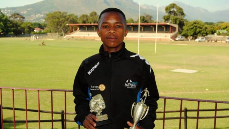 Former Young Bafana player Asanda Dyani standing in front of a sports field with two trophies in his hands.